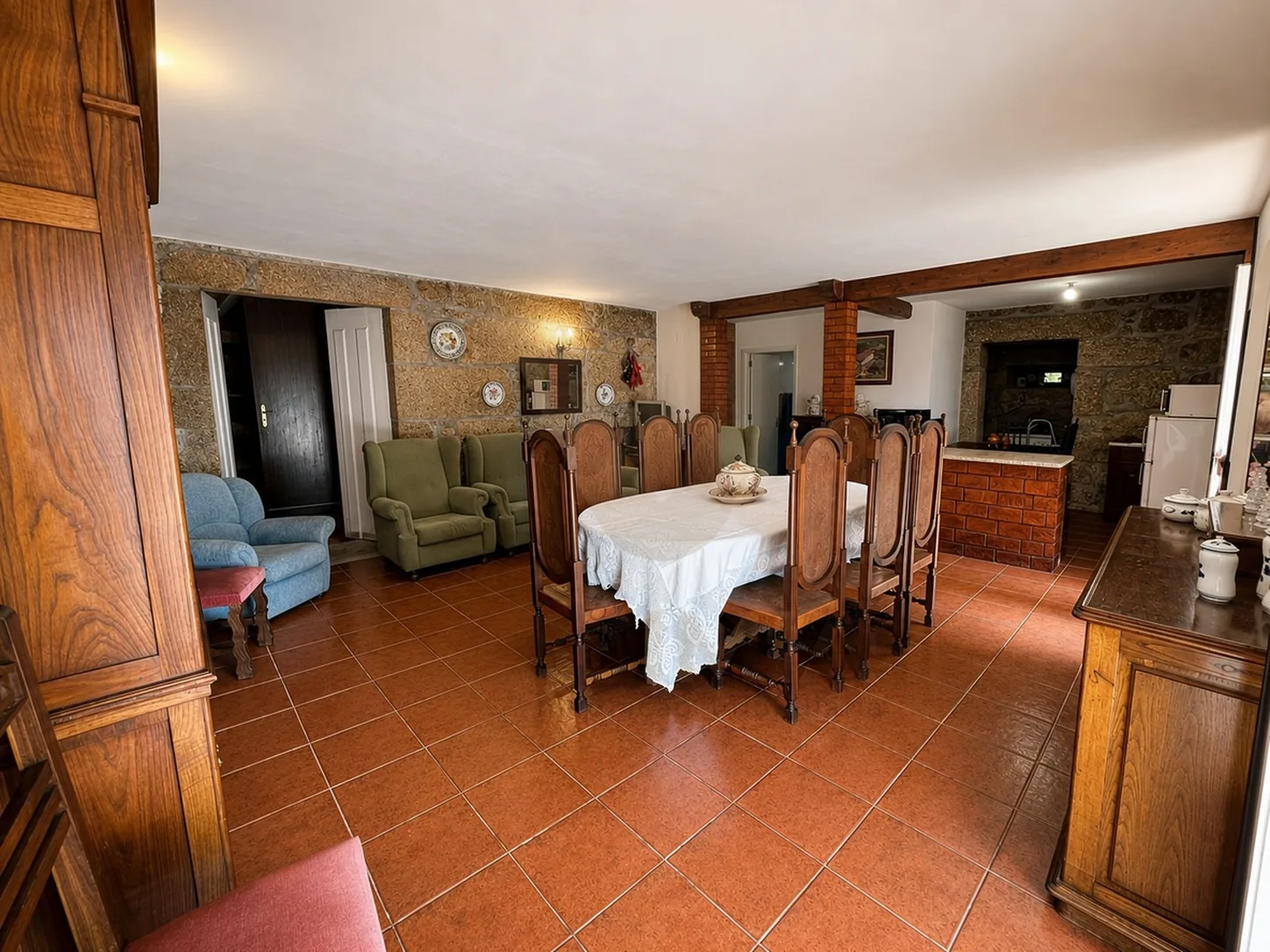 Dining and living room with a long table, carved chairs, and green and blue armchairs, opening onto a brick kitchen counter