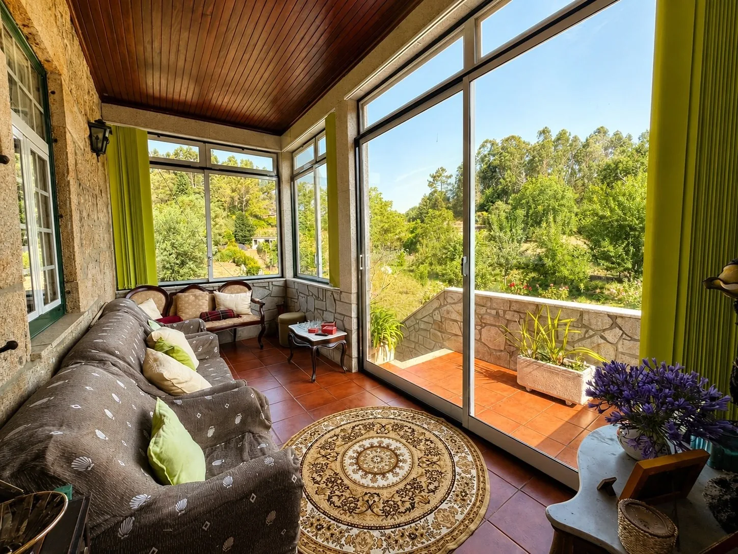 Glassed-in sunroom with a wood ceiling, green trim, a sofa, and floor-to-ceiling windows looking out over the trees and garden